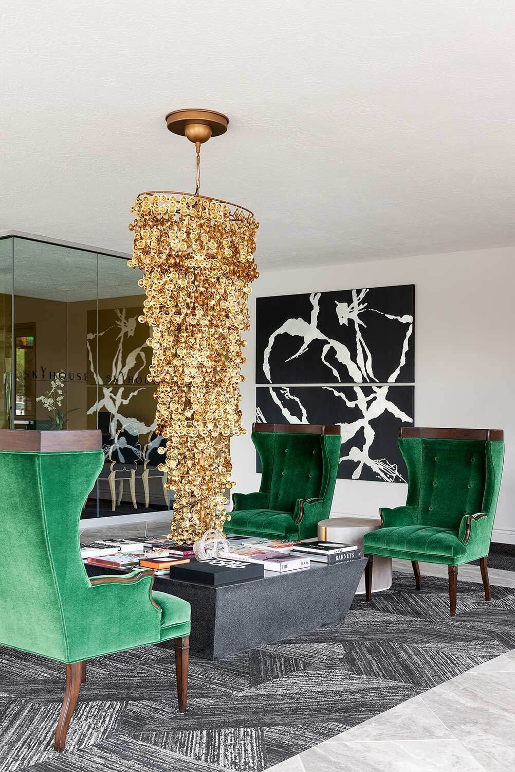 A modern and decorative foyer with emerald green arm chairs and a large gold chandlier with shingles. There is a coffee table that is filled with an assortment of magazines.